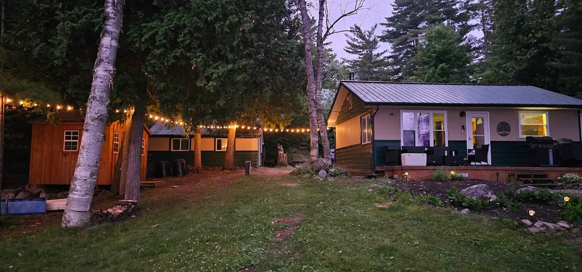 The cottage and the bunkie lit at dusk under string lights strung between birches